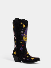 A black western-style cowboy boot with a pointed toe and low block heel, decorated with colorful sequined stars and a purple witch hat patch with a yellow jack-o'-lantern face, shown against a light gray background.