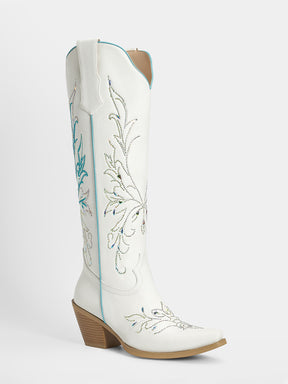 A white leather cowboy boot with turquoise and silver floral embroidery, a brown stacked heel, and a pointed toe, shown against a light gray background.