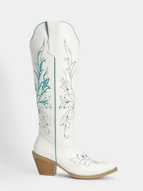 A white leather cowboy boot with turquoise and silver floral embroidery, a pointed toe, and a stacked wood-look block heel, shown against a light gray background.